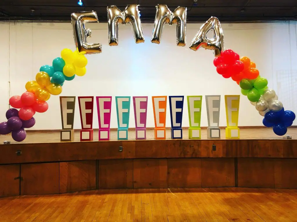 A beautiful arch made of Purple, Ruby Red, Gold, Green, Yellow, Red, Orange, Spring Green, Silver, and Dark Blue latex balloons, featuring Silver Letter "EMMA", perfect for adding a festive and personalized touch to an anniversary party in Brooklyn, delivered by Balloons Lane Balloon Delivery.