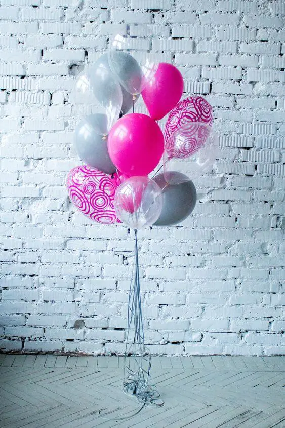 A combination of gray, bright pink, and clear balloons with printed mylar accents. The printed mylar adds a fun and whimsical touch to the design, making it a great addition to any celebratory event.