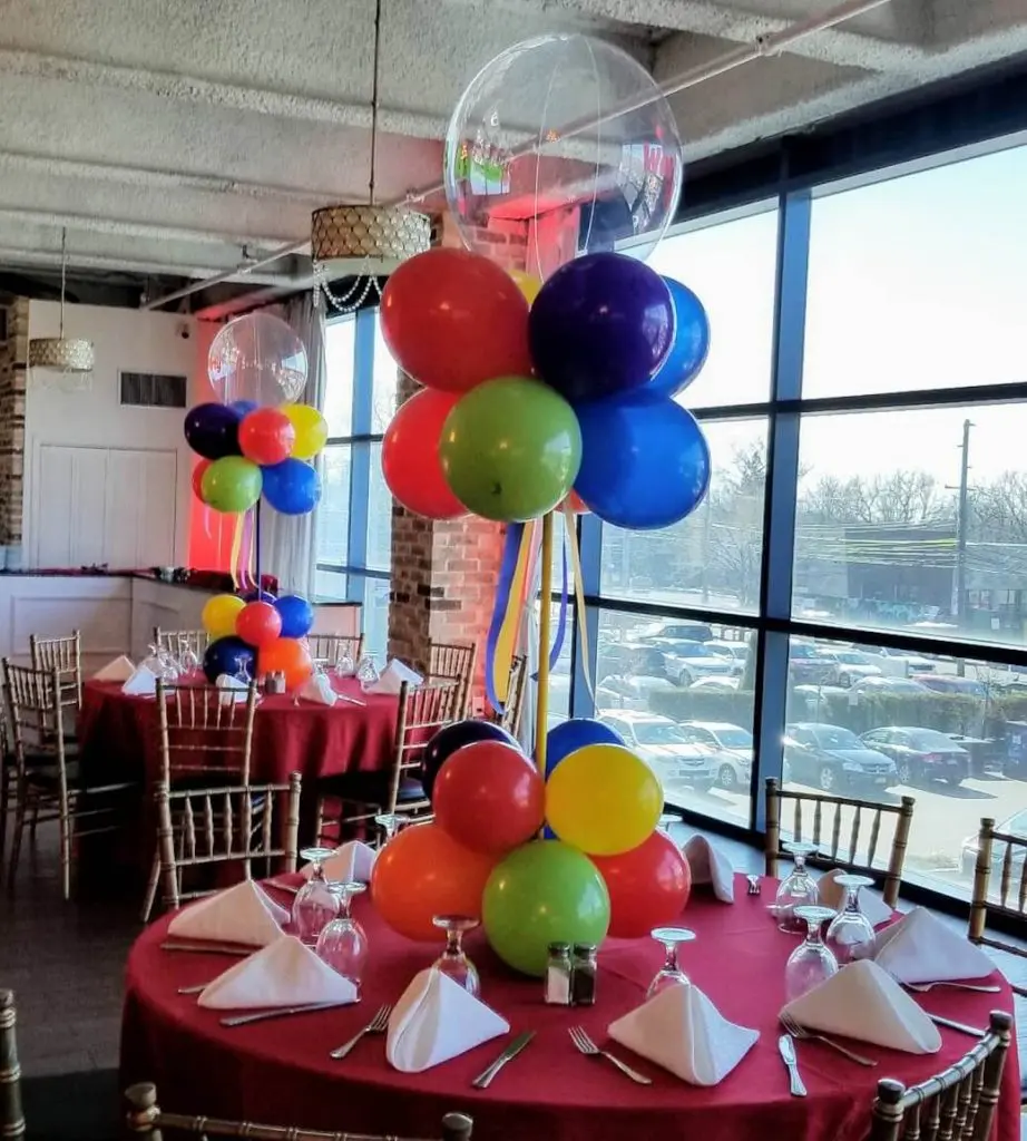 A fun and colorful decoration for a baby's first birthday party, featuring a mix of red, purple, blue, green, orange, yellow, and ruby red latex balloons arranged in a decorative manner. The centerpiece creates a festive and playful atmosphere for the celebration.