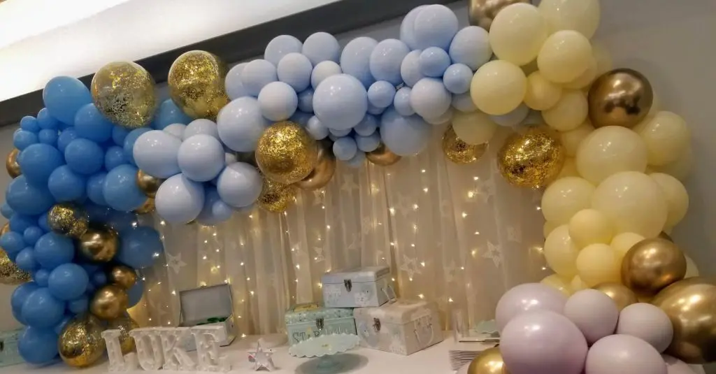Balloons Lane in Brooklyn showcases a pastel-colored balloon garland arch in shades of blue, gold, yellow, ivory, and pink, perfect for an anniversary celebration or a bar mitzvah. The balloons are arranged in an arch shape, creating an eye-catching display.