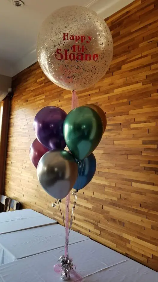 A stunning and festive decoration for an event or party, featuring a mix of Chrome Blue, Chrome Green, Chrome Silver, Chrome Gold, Chrome® Mauve, and Chrome Pink balloons. The centerpiece includes big round clear confetti balloons and personalized balloons, along with Chrome helium balloons arranged in a decorative manner.