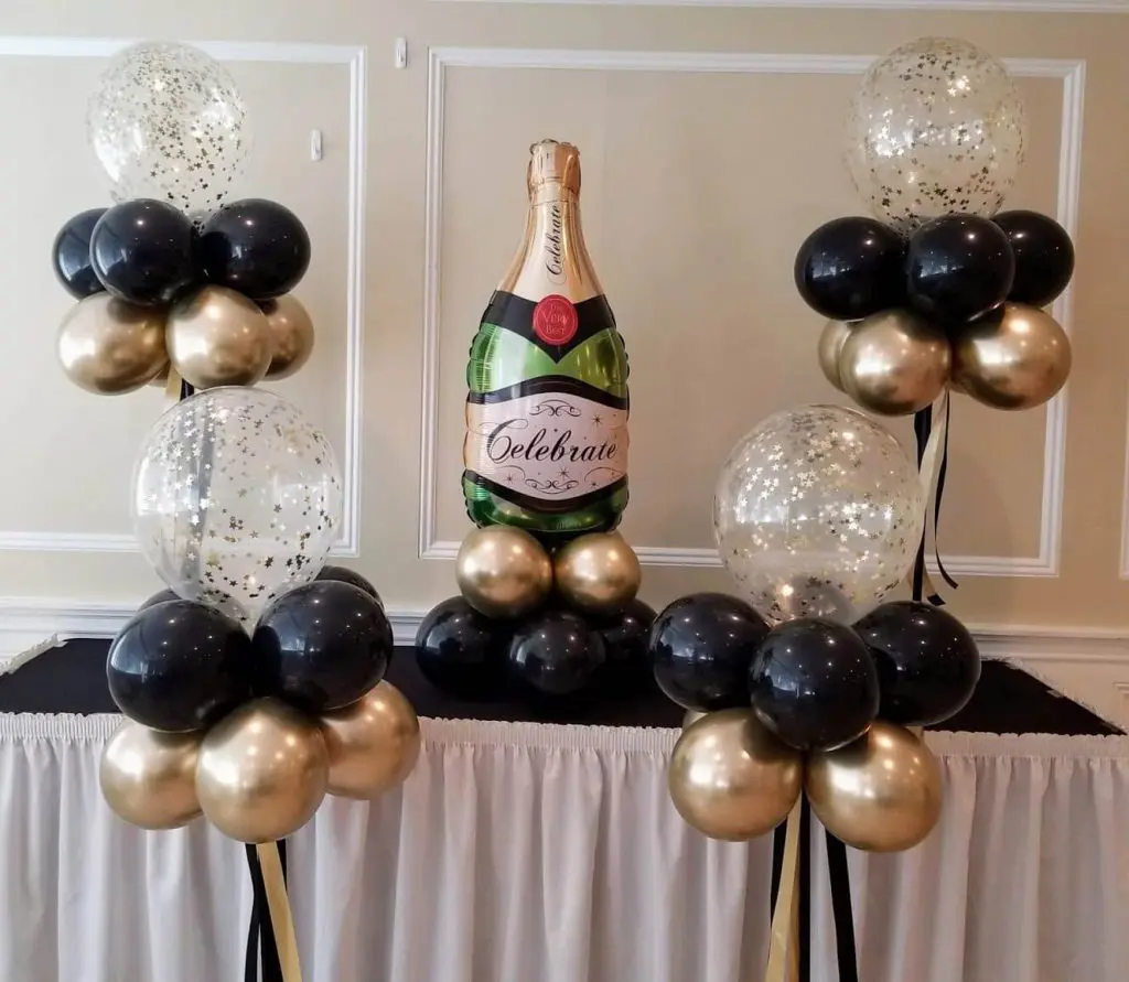 A glamorous Hollywood-themed balloon centerpiece created for a party on Balloons Lane in Brooklyn. The centerpiece features a mix of black, gold, and silver balloons, including confetti balloons and a large champagne bottle-shaped balloon. The balloons are arranged in a tall column and tied to a weight at the base, creating a dramatic and festive atmosphere for the occasion.