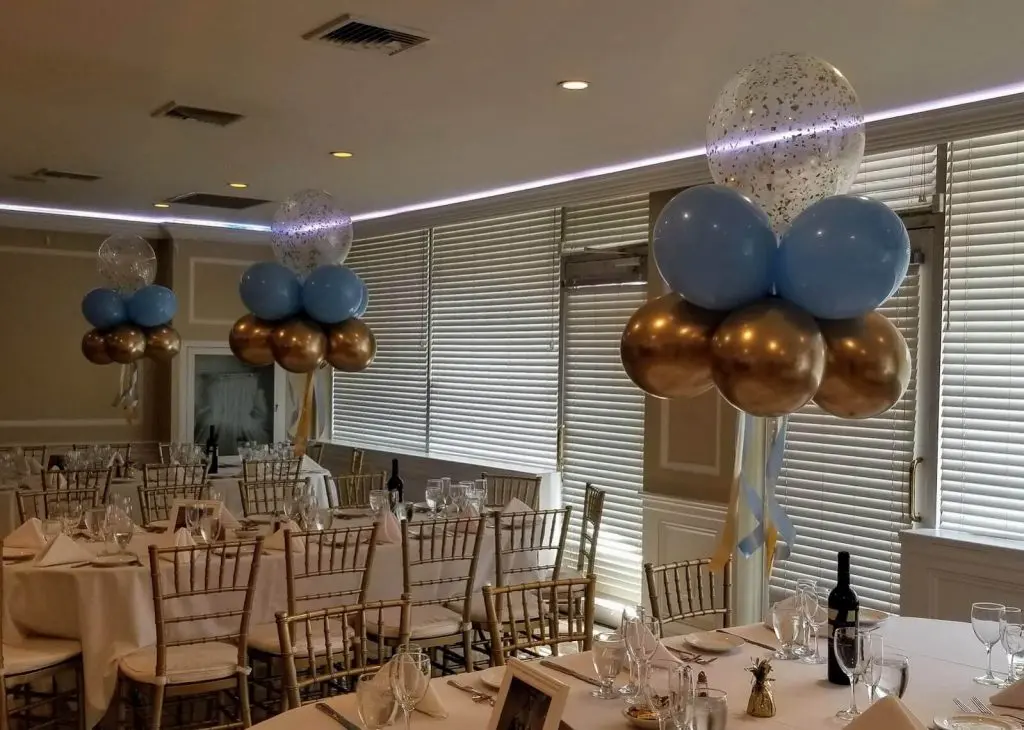A centerpiece stand with a clear 16-inch balloon filled with gold confetti, surrounded by light blue, chrome gold, Caribbean blue, and white balloons. This decoration is perfect for adding a touch of elegance and celebration to any party.