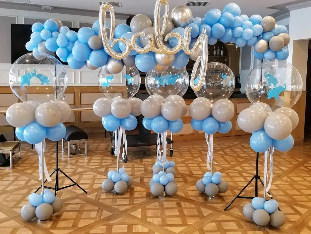 A baby shower balloon arrangement featuring light blue and gray latex balloons with an elephant theme. There is also a clear balloon included to make the arrangement stand out.