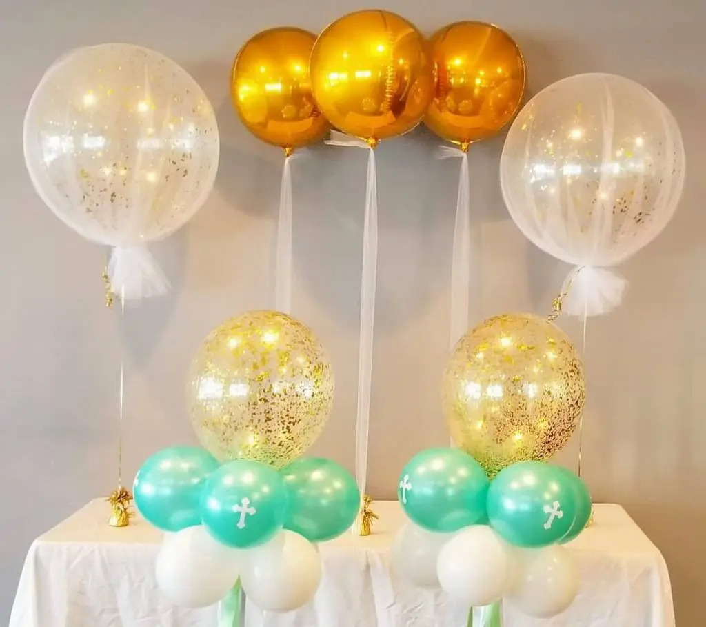 A winter-themed balloon decoration created on Balloons Lane in Staten Island. The centerpiece features a clear round tulle wrapped around a weight and filled with green and gold orbz balloons as well as confetti balloons. The balloons float above the centerpiece, adding a festive touch to the winter green and gold color scheme.