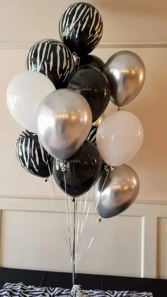 A bold and stylish balloon centerpiece in black, silver, white, and zebra print delivered to a location in NYC for a special occasion. The centerpiece features a mix of zebra print and solid black, silver, and white balloons arranged in a tall, dramatic column. Smaller black and white zebra print balloons are also scattered around the base of the centerpiece, adding to the festive atmosphere.