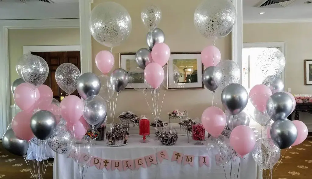A lovely christening table decoration featuring pink chrome, silver, and white balloons arranged on Balloons Lane in Brooklyn. The centerpiece includes a mix of large and small balloons tied to a weight and placed in the center of the table. The balloons create a beautiful and celebratory atmosphere for the decoration.