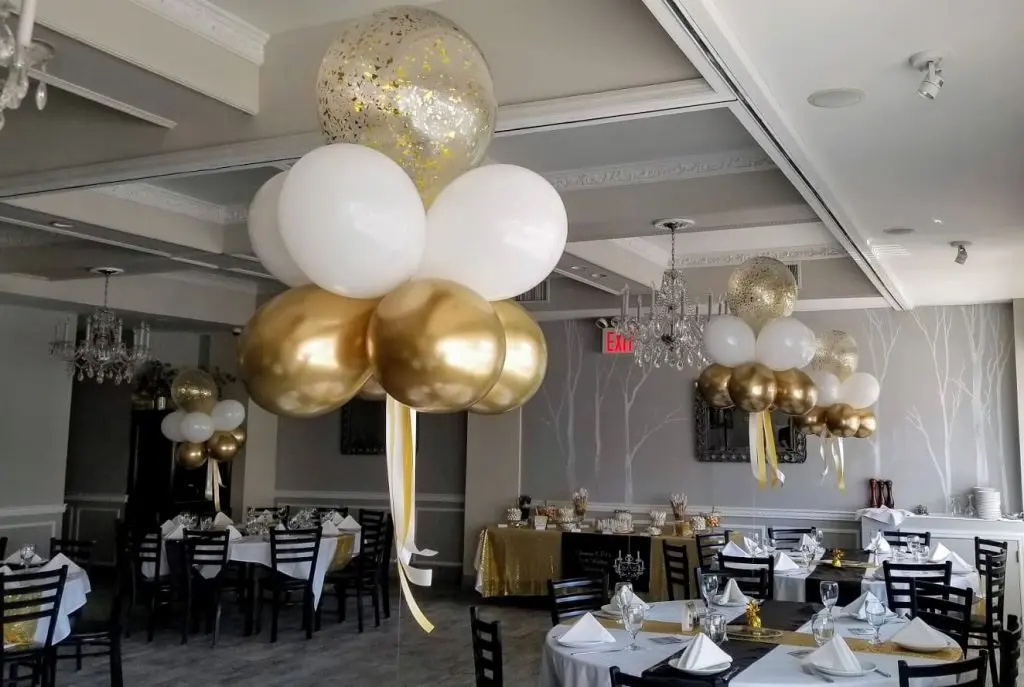 A stunning balloon centerpiece in gold and white confetti balloons created for an anniversary party decoration on Balloons Lane in NYC. The centerpiece includes a mix of large and small balloons tied to a weight and placed in the center of the table. The gold and white confetti adds a touch of elegance and sparkle to the balloon decorations, creating a festive and celebratory atmosphere for the anniversary party.