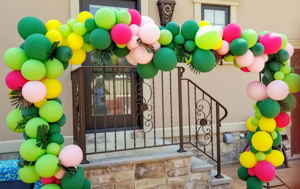 The balloons come in shades of emerald green, citrine yellow, wild berry pink, and pineapple. They are arranged into a garland arch, creating a cheerful and vibrant atmosphere.