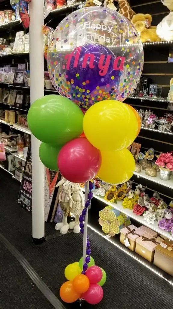 A colorful balloon tower created for an anniversary party on Balloons Lane in Brooklyn. The tower features a mix of pink, orange, lime green, yellow, and purple latex balloons and bubble balloons arranged in a spiral column. The balloons are tied to a weight at the base and create a festive and celebratory atmosphere for the party.