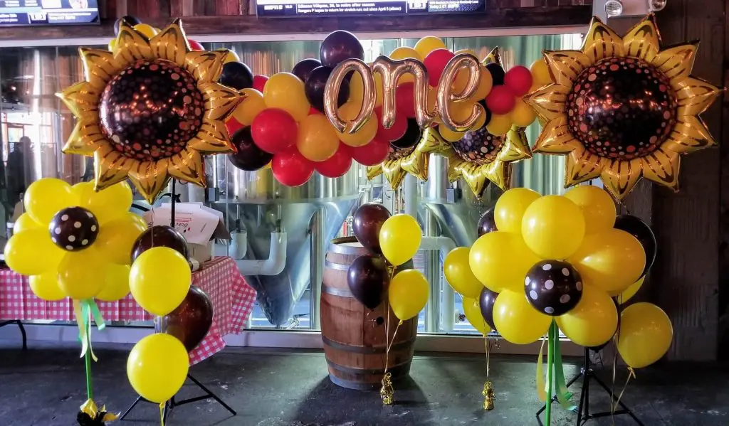 A garland arch made of balloons in a sunflower theme, created by Balloons Lane Balloon for a baby's first birthday. The arrangement features sunflower-shaped balloons in yellow and brown, with accents of green and white. A silver phrase balloon with the number "one" is included, adding a festive touch to the celebration.