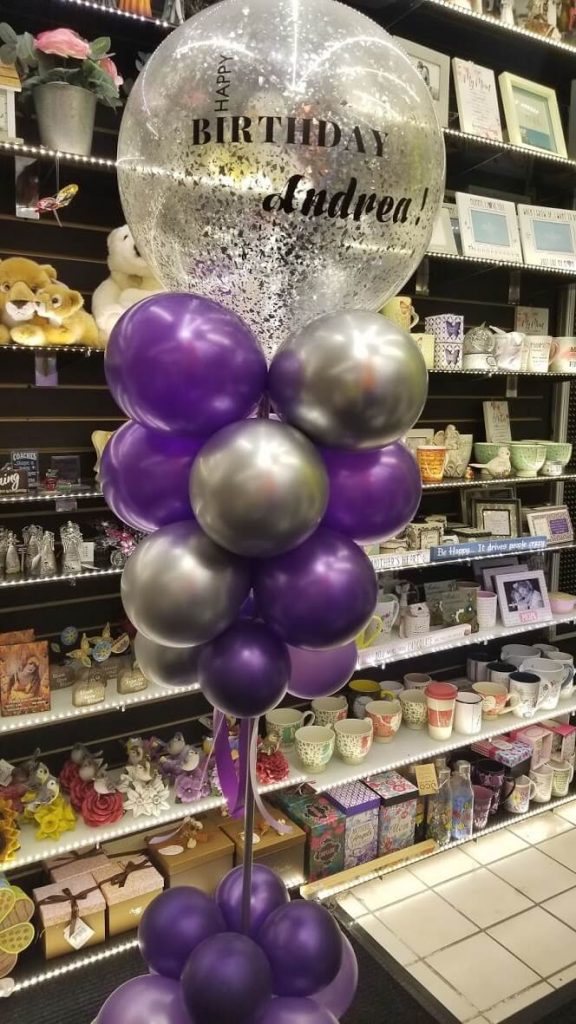 The centerpiece features jumbo purple and light purple balloons with silver confetti, as well as a personalized balloon with the girl's name. The balloons are tied to a weight at the base and arranged in a column, creating a fun and celebratory atmosphere for the occasion.