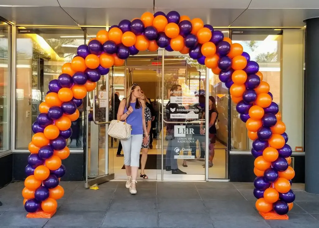 A balloon arch created by Balloons Lane in Brooklyn, featuring orange and purple balloons. The colors are bold and vibrant, creating a festive and energetic atmosphere suitable for an event.