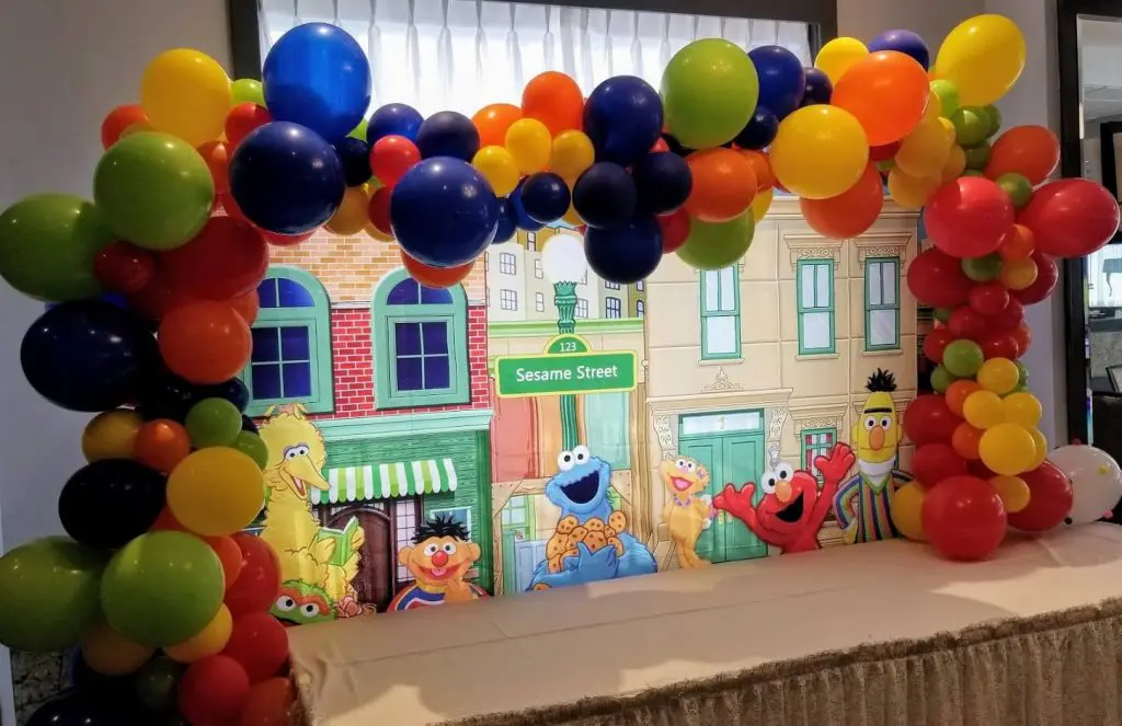 A balloon arch created by Balloons Lane in NJ, featuring green, purple, orange, yellow, red, and black balloons. The colors are bright and bold, creating a lively and colorful atmosphere suitable for a party.