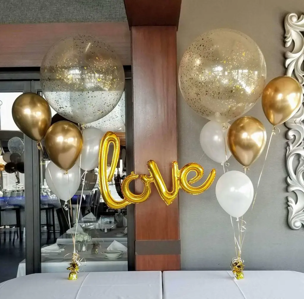 A Valentine's Day balloon display for a dessert table in New Jersey, featuring Pearl White, Chrome Gold, and White balloons, with gold confetti balloon bouquets and Love script mylar balloons.