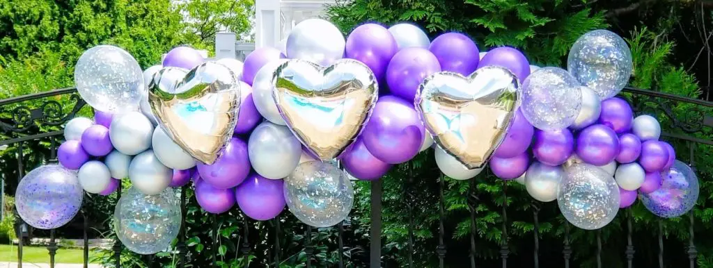 A Valentine's Day balloon decoration in Brooklyn, featuring a garland arch made of chrome silver, chrome purple, and white balloons, with big chrome silver heart balloons as the centerpiece.