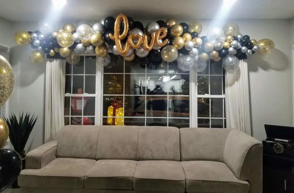 A Valentine's Day balloon garland in shades of chrome gold, chrome silver, chrome black, and gold, featuring Love balloons and some confetti balloons, displayed on a window railing for decor in Brooklyn.