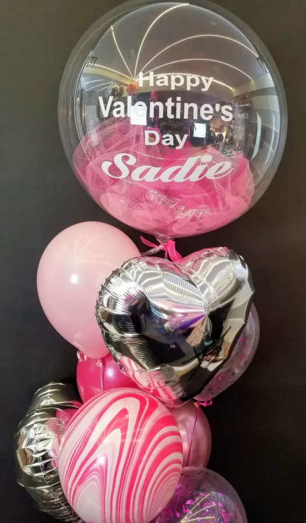 A Valentine's Day balloon bouquet in shades of purple, silver, light rose pink, hot pink, lavender, and purple, featuring big heart-shaped balloons and clear personalized bouquet balloons, in New Jersey.