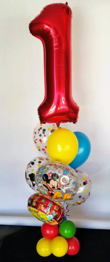 A cheerful balloon decoration featuring yellow, blue, white, and green balloons, with large red number "1" balloons as the centerpiece.