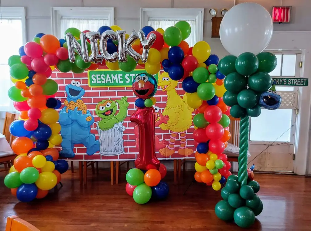 A festive balloon arrangement for a first birthday party in New York City, featuring a red number 1 balloon as the centerpiece, as well as a balloon bouquet, arch, garland, tree, and column. The display also includes confetti balloons and customized name balloons in silver.