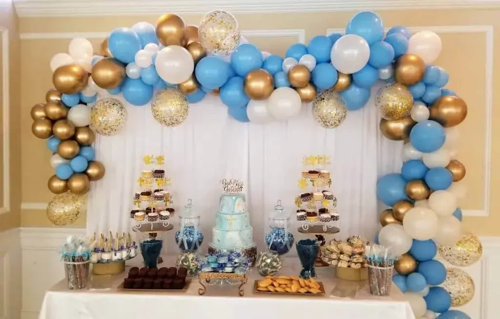 A gorgeous balloon garland arch made up of pearl white, light blue, chrome gold, and gold confetti balloons in different sizes, mounted on an arch frame covered in green foliage.