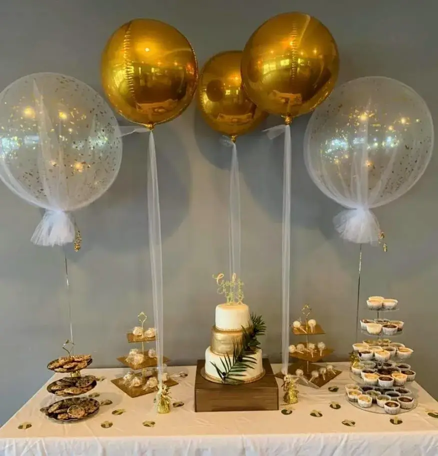 A large clear balloon filled with gold confetti and surrounded by smaller gold balloons, wrapped in a net, on a tea table.