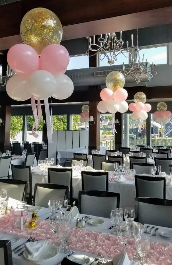 A beautiful balloon bouquet featuring light pink, baby pink, and white balloons, with gold confetti and pink ribbon bows, perfect for a special event centerpiece.