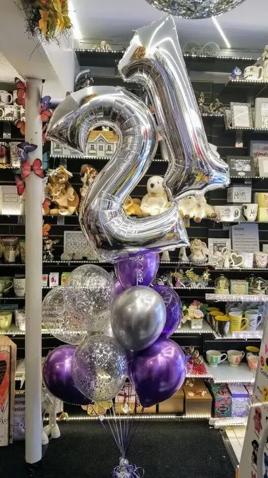 A festive balloon bouquet featuring purple, grey, and confetti balloons, with a large number "21" balloon and a letter balloon.