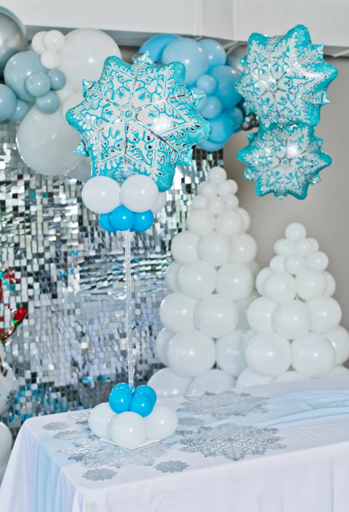 A Winter Wonderland balloon garland arch kit with large snowflake chrome blue, white, and silver balloons, suitable for Snow Queen Princess-themed girl's birthday parties or "Baby, It's Cold Outside" baby shower decorations. Additionally, a tabletop balloon tree adorned with shimmer wall snowflakes and funny faces.