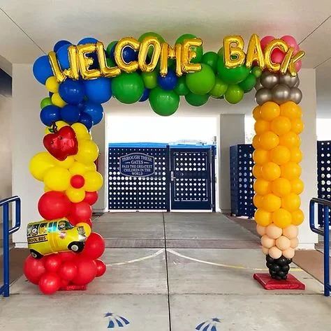 Mylar letter balloons spelling "WELCOME BACK" surrounded by red, blue, yellow, pink, and peach balloons, with additional red apple and car balloons.
