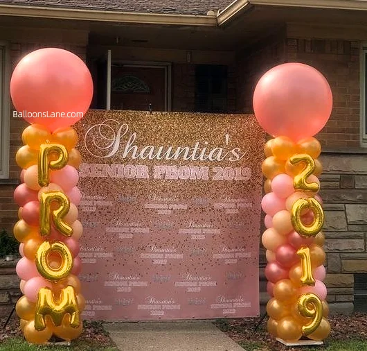 Graduation balloon column featuring a blend of peach pink, pink, orange, and gold latex balloons, adorned with "PROM" letter balloons and corresponding number balloons in matching colors.