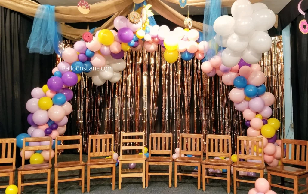 Pastel-colored balloon arch with purple, pink, lavender, yellow, and blue balloons, adorned with white balloon clusters and matching pastel-colored balloon bunches, perfect for celebrating a themed event in school.