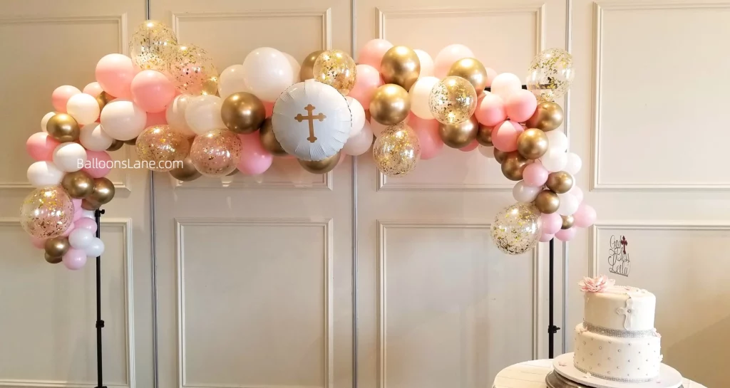 Balloon backdrop for christening featuring gold, pink, white, and confetti balloons, with a customized white Communion balloon in the center, in NJ.