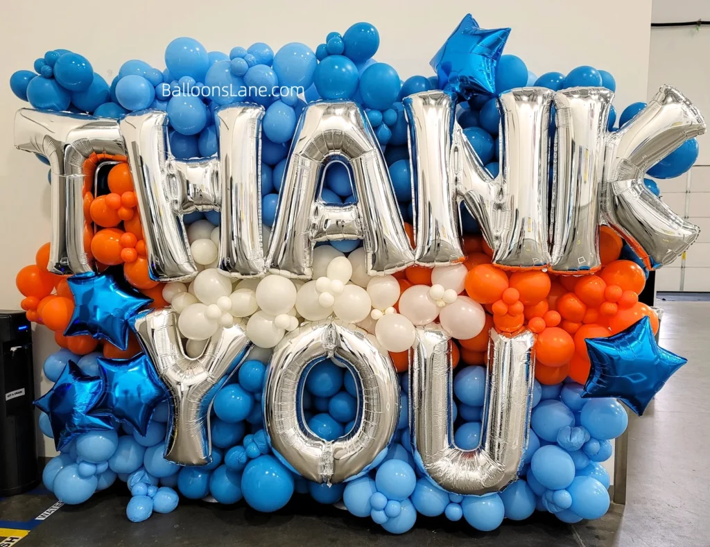 Giant balloons back drop with alrg letter balloons in silver blue latex and foil balloon and orange and white balloon used as abackdrop to say thank you