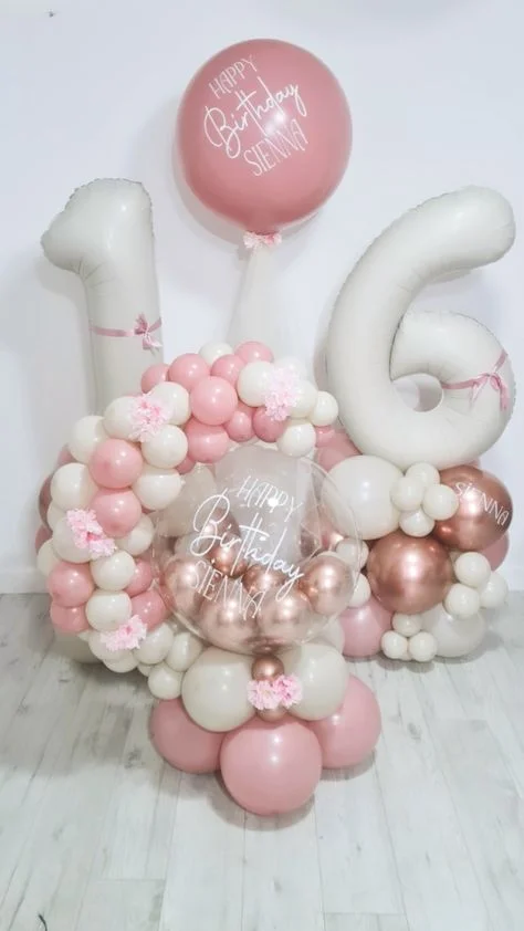 A display featuring white number balloons 1 & 6, along with customized birthday balloons in pink and white, and a bouquet of pink and rose gold balloons in NYC.