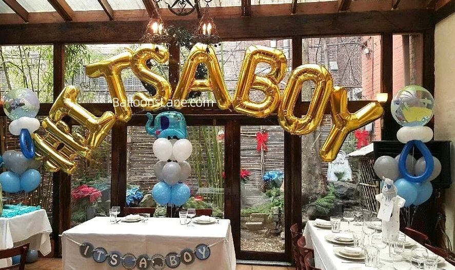 A gender reveal balloon arrangement featuring "It's a Boy" letter balloons, blue, white, and grey balloons, twisted blue balloons, and an elephant balloon.